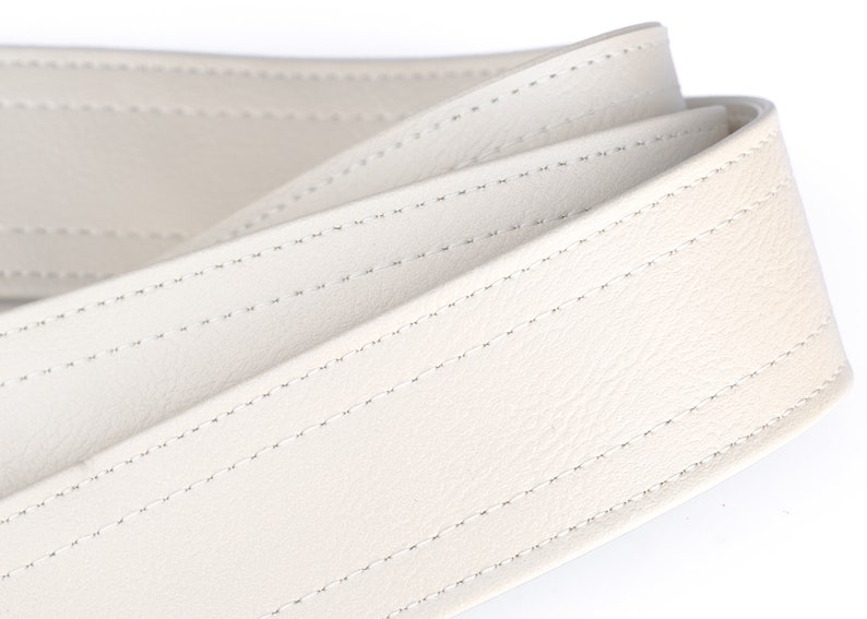May include: Close-up of a cream-colored leather belt. The belt is folded over itself, showcasing the detailed stitching. The stitching is a lighter color than the belt, creating a subtle contrast. The leather appears smooth and supple.