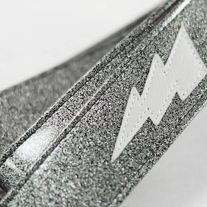 May include: A silver glitter guitar strap with a white lightning bolt design.