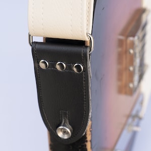 May include: Close-up of a guitar strap with a cream-colored shoulder pad and black leather end piece. The black leather end has three silver rivets and a silver button. The strap is attached to a sunburst-colored guitar.