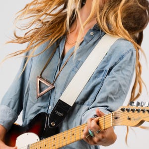 May include: A white guitar strap with black leather ends is worn over a denim shirt. The strap is attached to a sunburst electric guitar. The guitar has a maple neck and a Fender headstock. A triangular pendant hangs from a necklace.