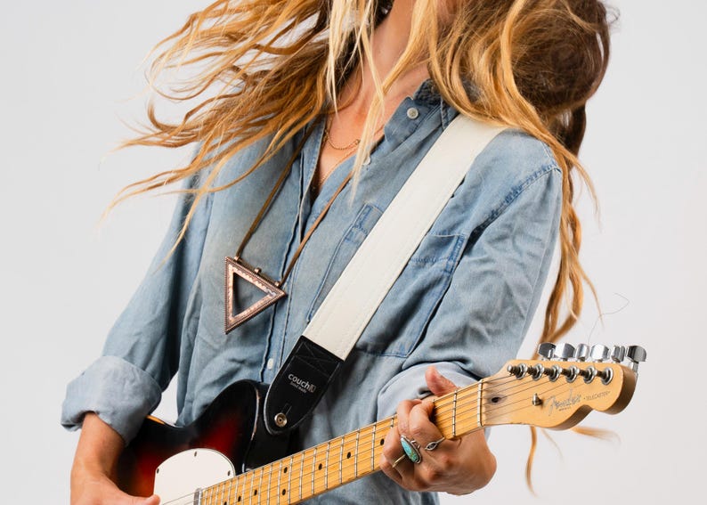 May include: A white guitar strap with black leather ends is worn over a denim shirt. The strap is attached to a sunburst electric guitar. The guitar has a maple neck and a Fender headstock. A triangular pendant hangs from a necklace.