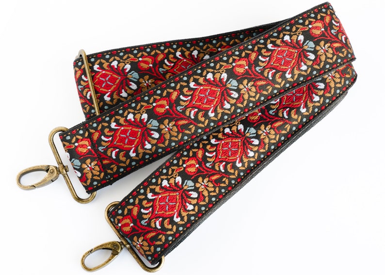 May include: A black and red patterned guitar strap with gold metal clasps. The strap has a floral design with red, gold, and white accents.