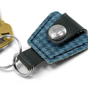 May include: A blue and black leather key chain with a silver button featuring a logo. The key chain is attached to a silver key ring.