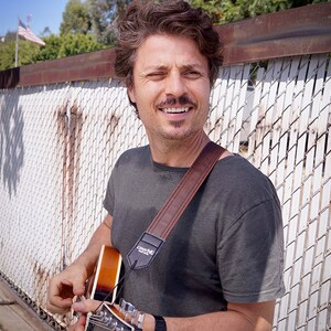 May include: A man with a mustache and a brown leather guitar strap smiles while holding a guitar. The strap has the word "Couch" printed on it.