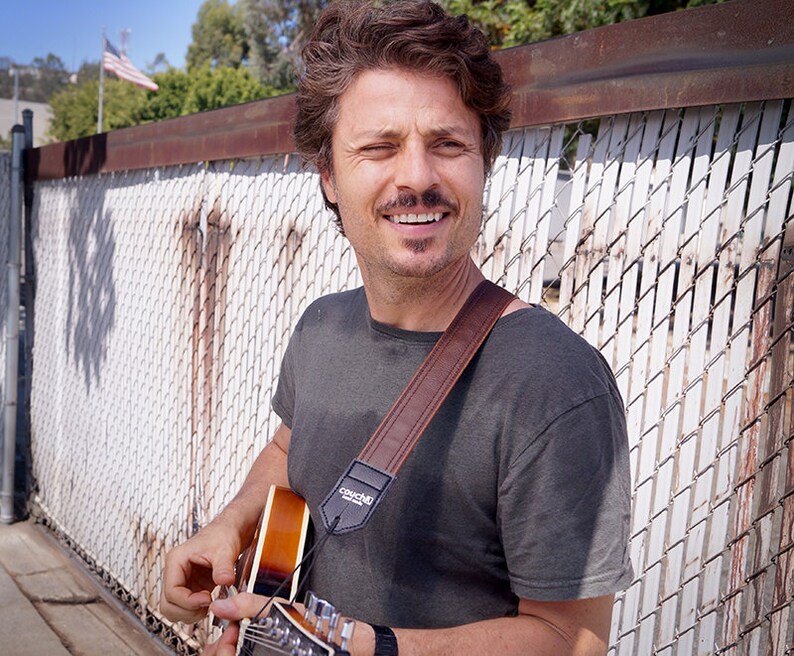 May include: A man with a mustache and a brown leather guitar strap smiles while holding a guitar. The strap has the word "Couch" printed on it.