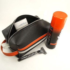 May include: Black leather travel toiletry bag with orange and white stripes. The bag is open and contains a white jar of face cream and a razor. A can of shaving cream is next to the bag.