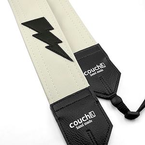 May include: A white leather camera strap with a black lightning bolt design. The strap has black leather ends with the text "couch[ ]hand made" on each end.