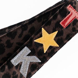 May include: A brown and black leopard print strap with a black leather accent. The strap features a silver letter "K", a yellow star, and a red letter "T" applique.