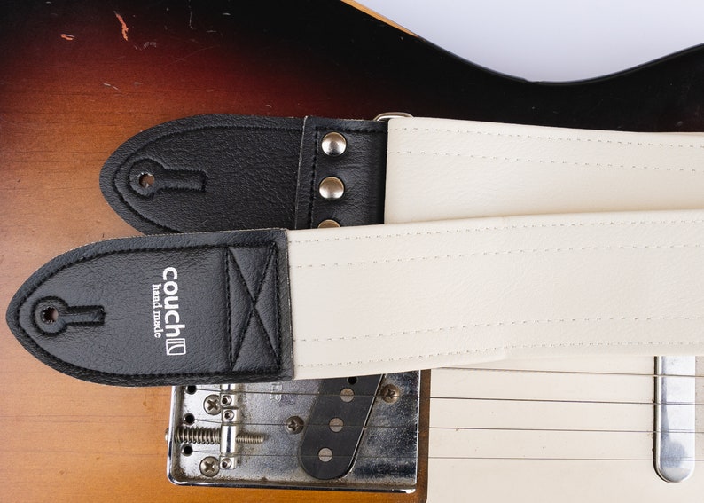 May include: Two guitar straps, one black and one cream, are shown against a guitar. The black strap has a black leather end with the text "couch hand made". The cream strap has a cream leather end. Both straps are made of a sturdy material.