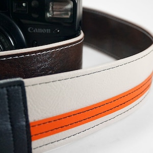 May include: A white camera strap with a brown leather section and an orange stripe. The strap is stitched with white thread. The strap is designed to be used with a camera.