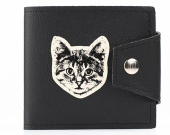May include: Black bi-fold wallet with a cat face graphic. The cat face is cream-colored with black markings. The wallet has a snap closure with a silver button. The wallet is made of a textured material.