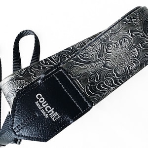 May include: A black and silver camera strap with a floral embossed design. The strap has a black leather patch with the text "couch" and "hand made" in white.