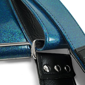 Pu&ograve; includere: Primo piano di una tracolla per chitarra blu glitterata con una tracolla in pelle nera e una fibbia argentata. La tracolla ha una fibbia argentata con la parola "Cosmic" incisa.