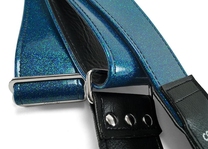 Pu&ograve; includere: Primo piano di una tracolla per chitarra blu glitterata con una tracolla in pelle nera e una fibbia argentata. La tracolla ha una fibbia argentata con la parola "Cosmic" incisa.