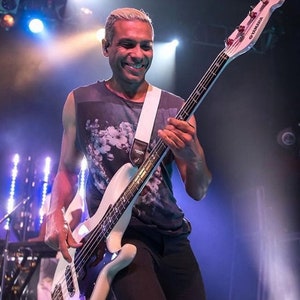 May include: A musician playing a white Yamaha bass guitar on stage. The bass has a white body, a black fretboard, and a white strap. The musician is wearing a dark sleeveless shirt with a floral design.