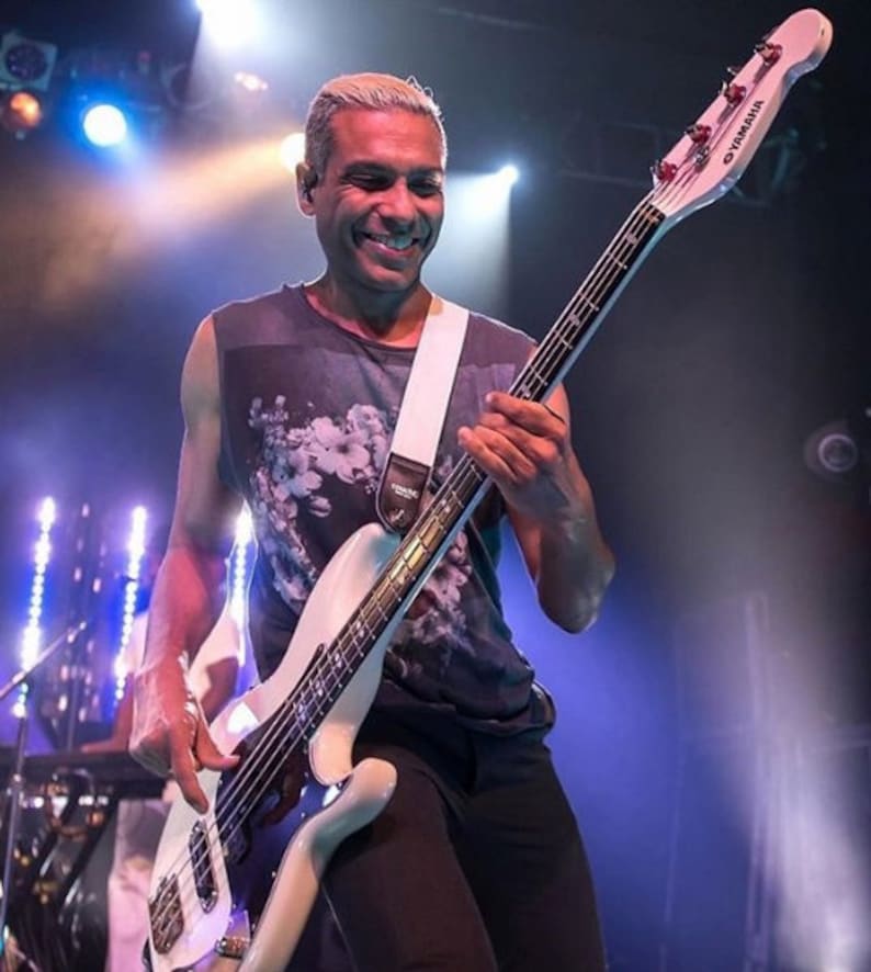 May include: A musician playing a white Yamaha bass guitar on stage. The bass has a white body, a black fretboard, and a white strap. The musician is wearing a dark sleeveless shirt with a floral design.