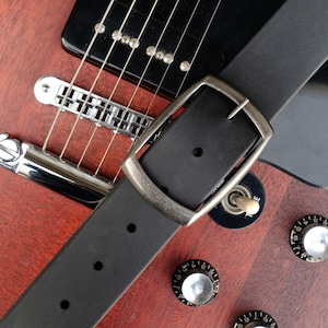 May include: A black leather guitar strap with a silver buckle. The strap is on top of a red electric guitar.