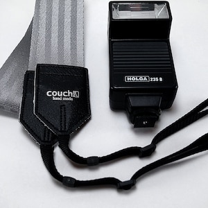 May include: A black camera flash with the text "HOLGA 235 B" on it. The flash is attached to a black camera strap with a gray and white striped camera strap.