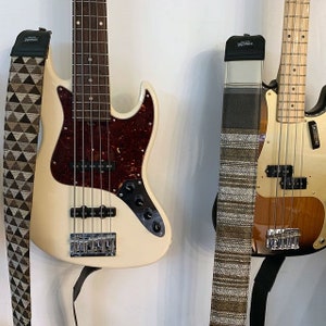 Puede incluir: Dos bajos el&eacute;ctricos con cuerpos y vetas de madera de diferentes colores. Uno tiene un cuerpo blanco con un m&aacute;stil de madera marr&oacute;n y una correa de guitarra con estampado marr&oacute;n y negro. El otro tiene un cuerpo marr&oacute;n con un m&aacute;stil de madera marr&oacute;n claro y una correa de guitarra con estampado marr&oacute;n y blanco.