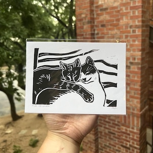 Puede incluir: Grabado en linóleo en blanco y negro que representa a dos gatos abrazándose. La impresión se titula "Fur Friend". La obra de arte se muestra en una funda de plástico transparente.