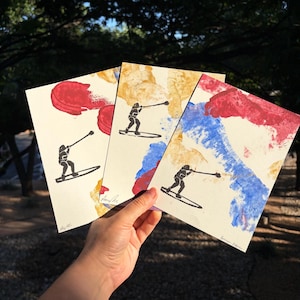 Può includere: Tre stampe artistiche rettangolari su carta color bianco sporco, ognuna con la silhouette nera di una persona su una tavola da paddle che usa un lungo bastone. Le stampe presentano pennellate di colore rosso, blu e giallo sulla superficie.