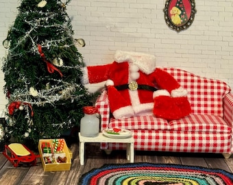 May include: Miniature Christmas scene with a decorated tree, Santa suit on a red and white checkered couch, and a colorful rug. A small table holds a jar and plate. A framed picture hangs on the white brick wall.