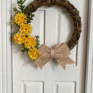 May include: A miniature circular wreath constructed from twisted brown paper vine, featuring a cluster of small yellow flowers with green foliage on the left side and a textured tan burlap bow centered at the bottom of the wreath frame.