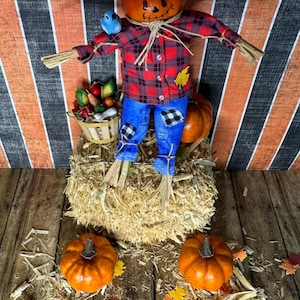 May include: A scarecrow figurine with a pumpkin head, wearing a red and black plaid shirt and blue pants with patches, stands on a hay bale. A small blue bird sits on its shoulder, and a basket of miniature vegetables rests beside it on the hay.