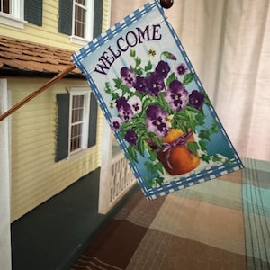 May include: A decorative welcome flag featuring a floral design with purple pansies and a peach-colored pot. The flag has a blue and white checkered border and the word "WELCOME" in white lettering. The flag is attached to a wooden pole.