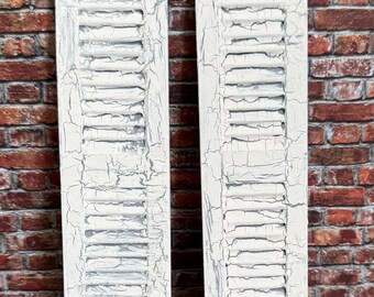 May include: Two distressed white wooden shutters with horizontal slats. The shutters have a weathered, crackled paint finish, giving them a vintage or farmhouse aesthetic. They are displayed against a brick wall.