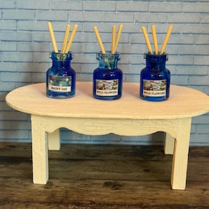 May include: Three small, cobalt blue glass bottles with wooden reeds, labeled "Rainy Day" and "Wild Flowers", sit on a cream-colored, oval table. The table has four legs and is set against a light blue brick wall. The bottles are likely miniature home decor items.