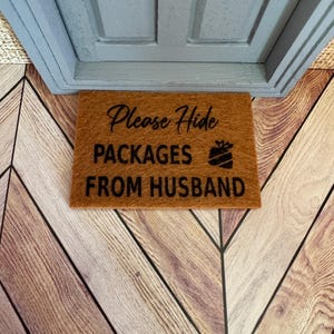 May include: A brown doormat with the text "Please Hide Packages From Husband" and a small gift box icon.