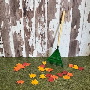 May include: A green plastic rake with a wooden handle rests on a bed of green moss, surrounded by colorful autumn leaves in shades of red, orange, and yellow. The background features a distressed, weathered wooden wall.