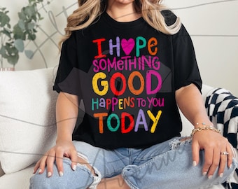 May include: Black t-shirt with the text "I hope something good happens to you today" in colorful letters. The shirt is worn by a person sitting on a white couch, wearing blue jeans.