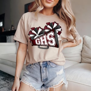 May include: A tan t-shirt featuring a maroon and white pom-pom design and a megaphone with the letters "GHS" in white. The shirt is made of a soft material and is suitable for casual wear.