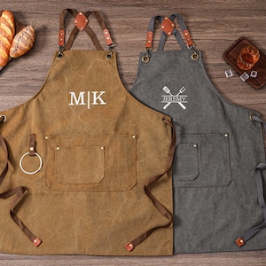 May include: Two aprons, one brown and one gray, with leather straps and pockets. The brown apron has the initials "MK" embroidered on it, while the gray apron has a crossed spatula and fork design with the name "Jeremy". Both aprons are designed for cooking or grilling.