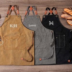 May include: Three personalized aprons in brown, gray, and black, with leather straps and pockets. The brown apron has the initials "K H", the gray apron has the name "Leroy", and the black apron has a crossed fork and knife design with the name "Jeremy".