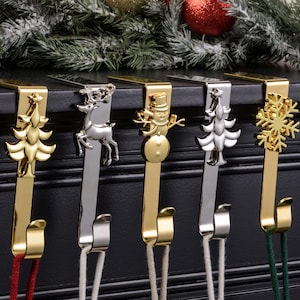 Op de afbeelding: Een set van vijf kerstsokkenhouders met feestelijke ontwerpen. Elke houder heeft een ander ornament met een vakantiethema: een kerstboom, een rendier, een sneeuwpop, een andere kerstboom en een sneeuwvlok. De houders zijn goud en zilver.