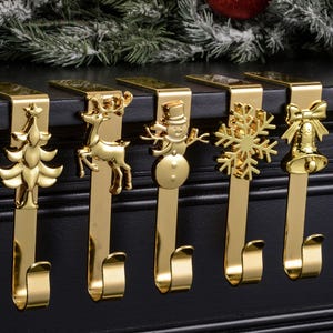 Può includere: Set di cinque ganci dorati per calze natalizie. Ogni gancio presenta un design a tema festivo diverso: un albero di Natale, una renna, un pupazzo di neve, un fiocco di neve e una campana con un fiocco. I ganci sono progettati per essere appesi a un camino.