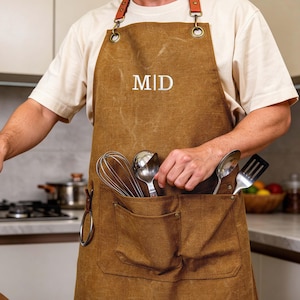 May include: Brown apron with leather straps and metal hardware. The apron has two pockets and the initials "MID" embroidered on the front. A spatula is held in one hand, while cooking utensils are in the pocket.
