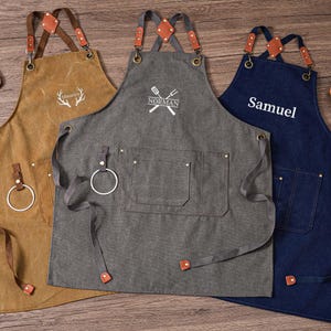 May include: Three aprons in brown, gray, and navy blue with leather straps and accents. The brown apron has a deer antler design, the gray apron has a crossed spatula and fork design with the name "Norman", and the navy blue apron has the name "Samuel".