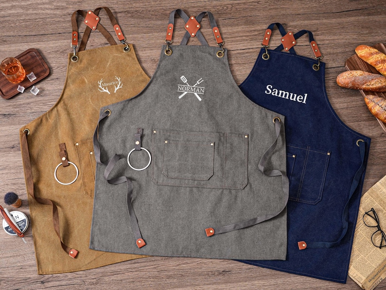 May include: Three aprons in brown, gray, and navy blue with leather straps and accents. The brown apron has a deer antler design, the gray apron has a crossed spatula and fork design with the name "Norman", and the navy blue apron has the name "Samuel".