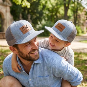 May include: Two light blue trucker caps with brown leather patches. One cap reads "Dad" and the other reads "Ethan". The caps have a mesh back and a curved peak. The caps are worn by a father and son.