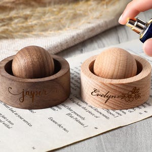 May include: Two wooden perfume bottle holders, one dark and one light, each with a wooden sphere. The dark holder is engraved with "Jasper", the light one with "Evelyn" and a floral design. A hand is spraying perfume.