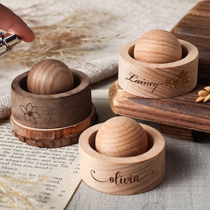 Puede incluir: Juego de difusores de aromaterapia de madera con tres recipientes redondos de madera, cada uno con una esfera de madera. Dos recipientes tienen nombres grabados, "Lainey" y "Olivia". Un recipiente tiene un diseño floral. Una mano rocía perfume.
