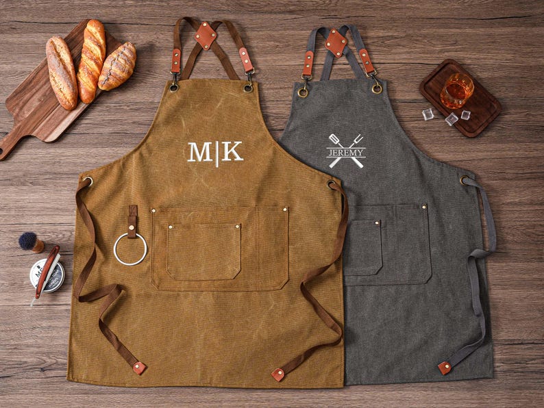 May include: Two aprons, one brown and one gray, with leather straps and pockets. The brown apron has the initials "MK" embroidered on it, while the gray apron has a crossed spatula and fork design with the name "Jeremy". Both aprons are designed for cooking or grilling.