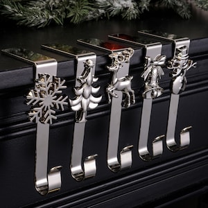 May include: A set of five silver Christmas stocking holders. Each has a different festive design: a snowflake, Christmas tree, reindeer, bow with bells, and a snowman. The hooks are designed to hang on a mantelpiece.