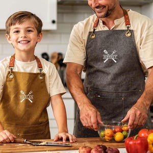 May include: A father and child wearing personalized aprons in a kitchen setting. The aprons are brown and gray, with leather straps and the names "Harlan" and "Jeffrey" printed on them, along with a crossed spatula and fork design. The scene includes fresh produce.