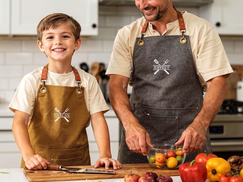 May include: A father and child wearing personalized aprons in a kitchen setting. The aprons are brown and gray, with leather straps and the names "Harlan" and "Jeffrey" printed on them, along with a crossed spatula and fork design. The scene includes fresh produce.
