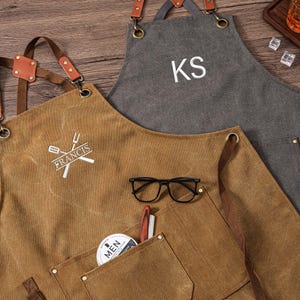 May include: Two aprons, one brown and one gray, with leather straps and pockets. The brown apron has the name "FRANCIS" and a crossed fork and spatula logo. The gray apron is personalized with the initials "KS". Black eyeglasses and a pen rest in the brown apron pocket.